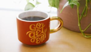 Steaming cup of coffee in a red mug with encouraging message "go get em" next to a green plant.